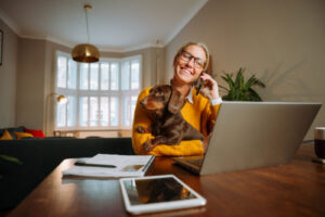 woman working on her laptop while holding her dachshund dog and talking on the phone at home